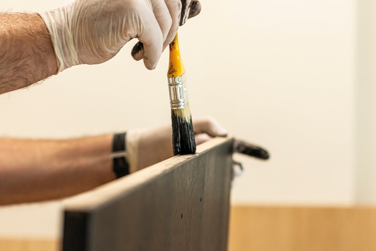 Person oiling a wooden surface with a brush, wearing gloves.