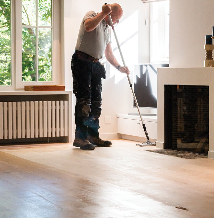 Person cleaning a wooden floor with a pad in a room with a fireplace and large window.